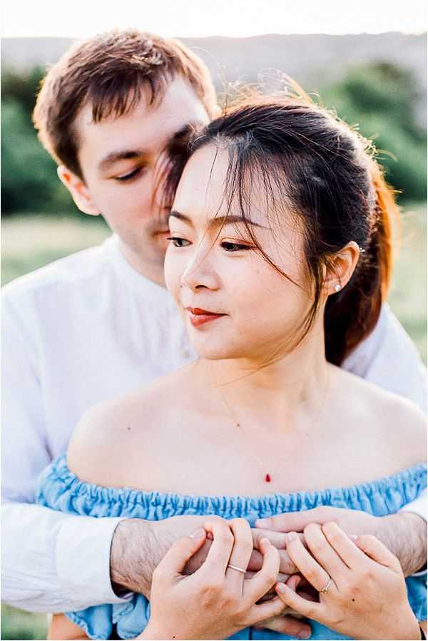 A close-up portrait of a couple outdoors, shot during golden hour with warm, soft light. The woman wears a light blue off-shoulder ruched top and a delicate red drop pendant necklace with small stud earrings, her dark hair pulled back in a loose updo with face-framing strands; she gazes softly to the side. The man, wearing a white long-sleeve shirt, stands behind her with his arms wrapped around her, their hands intertwined, both wearing rings. The composition is a tight portrait crop with a blurred green outdoor background, conveying an intimate, relaxed mood suited to an engagement or pre-wedding shoot.