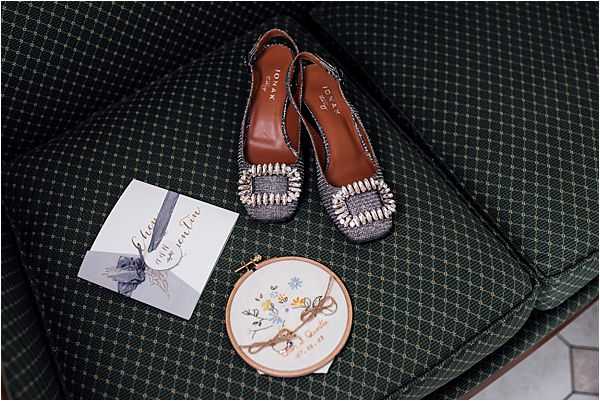 A flat lay detail shot arranged on a dark green diamond-patterned upholstered sofa or chair. The items displayed include a pair of Iotan brand slingback flats in a dark tweed fabric with large crystal and pearl embellished square buckles and tan leather insoles, a small embroidery hoop featuring hand-stitched floral and bird motifs with what appears to be a wedding date and names, and a wedding stationery card with a grey ribbon or bow detail. The composition is overhead and close-up, capturing bridal accessories and keepsakes as part of a getting-ready flat lay.