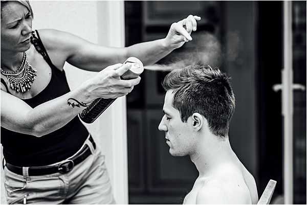 A black-and-white getting-ready photograph showing a hairstylist applying hairspray to a shirtless young man, likely the groom, who is seated on a wooden chair in profile view. The stylist, wearing a black tank top, light-colored pants, and a statement necklace, holds a comb in one hand while spraying with the other, with visible spray mist captured mid-air. The setting appears to be semi-outdoor or on a covered terrace, with a door visible in the background. The image is a close-up candid shot with strong contrast between light and dark tones.