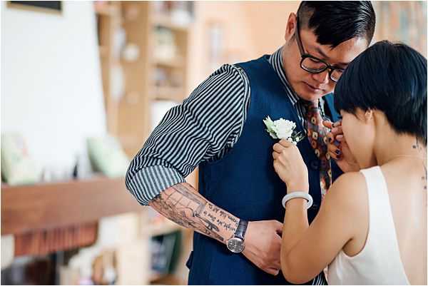 A close-up portrait of a bride pinning a small white boutonniere onto a groom's navy blue suit vest in what appears to be an indoor setting with bookshelves visible in the soft-focus background. The groom wears black-rimmed glasses, a navy vest over a blue and white vertical-striped shirt, and a patterned tie, with visible sleeve tattoos on his forearms and a watch on his wrist. The bride, with short dark hair, wears a sleeveless white top and a white watch, and leans in closely as she fastens the boutonniere. The styling has a relaxed, contemporary feel with personal, eclectic fashion choices rather than traditional formalwear.