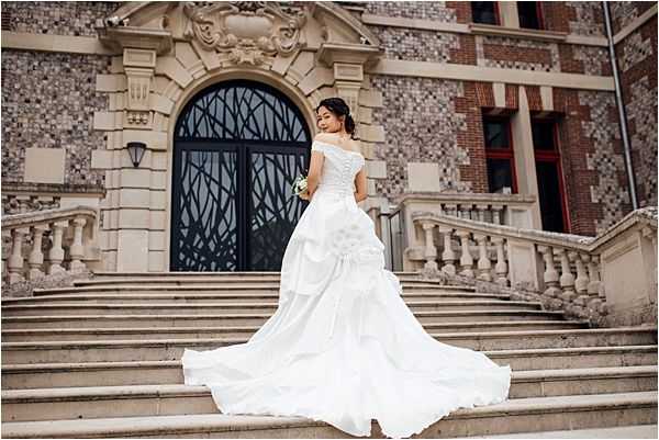 A bridal portrait taken outdoors on the grand stone staircase of a chateau-style building, with the bride posed with her back to the camera. She wears an off-the-shoulder white ball gown with a heavily ruffled skirt, corset-style lace bodice, and an extended cathedral train spread across the steps below her. She holds a small bouquet of white and green flowers at her side. The architecture behind her features an ornate arched doorway with decorative ironwork, flanked by a balustraded staircase railing. The overall styling is classic and formal. Wide portrait shot. Potential venue feature image.