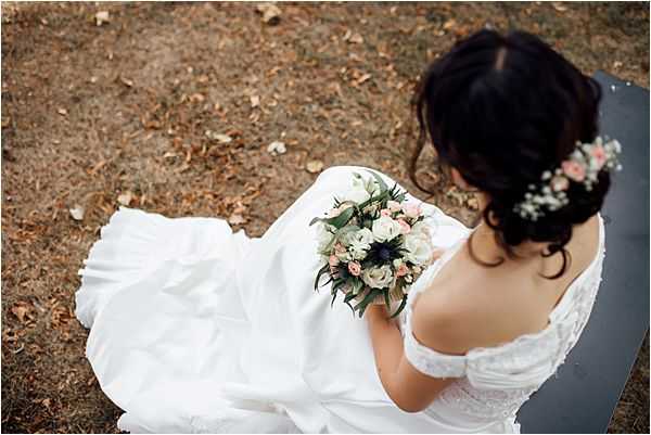 A bride is seated on a bench outdoors, photographed from an elevated overhead angle looking down. She wears an off-the-shoulder white ball gown with a full skirt that fans out around her. Her dark hair is styled in an updo adorned with small white and blush floral hair accessories. She holds a compact round bouquet featuring white ranunculus, blush pink roses, a deep navy-blue thistle, and green eucalyptus foliage. The composition is a portrait-style overhead shot that emphasizes the spread of her dress against the ground below.