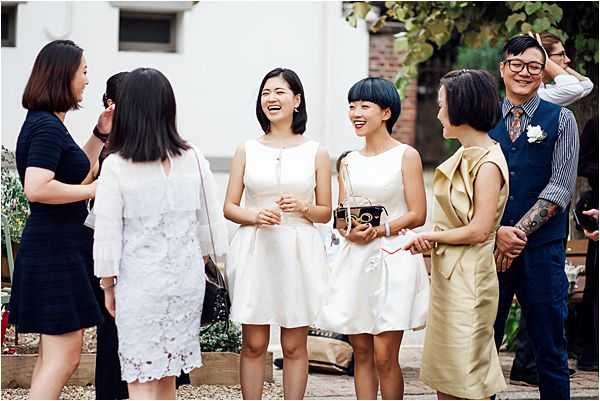 A candid group shot of approximately six guests socializing outdoors, likely during a cocktail hour or reception. The setting appears to be a courtyard or garden area with a brick and white building visible in the background. Two women in the center wear short white sleeveless dresses, one carrying a small vintage-style camera bag; nearby guests wear a white lace dress, a navy dress, a gold/champagne structured dress, and a man in a navy suit with a floral boutonniere. The group is laughing and engaged in conversation, captured in a relaxed, candid style at mid-range distance.