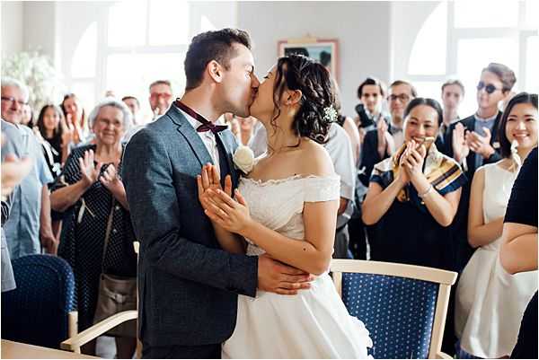 The bride and groom share their first kiss during an indoor civil ceremony, surrounded by approximately 20 guests who are clapping and smiling. The setting appears to be a French town hall (mairie), with bright white walls and large arched windows. The bride wears a white off-the-shoulder short-sleeve dress with her dark hair up, adorned with a small white floral hair piece, while the groom wears a grey suit with a burgundy bow tie and a white boutonnière. The shot is a medium portrait framing the couple in the center aisle with guests on either side, captured in a bright, airy, natural-light style.