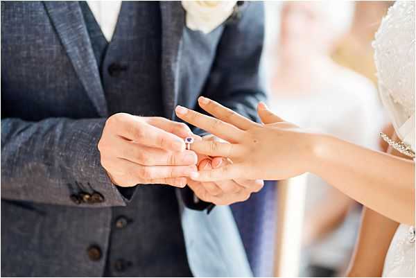 Close-up detail shot of the ring exchange moment during a wedding ceremony, showing the groom placing a ring with a small purple gemstone onto the bride's finger. The groom is wearing a navy blue three-piece suit with a white floral boutonniere, while the bride wears a lace-detailed white gown with a delicate bracelet visible on her wrist. The composition is tightly framed on the couple's hands, with the torsos softly blurred in the background.