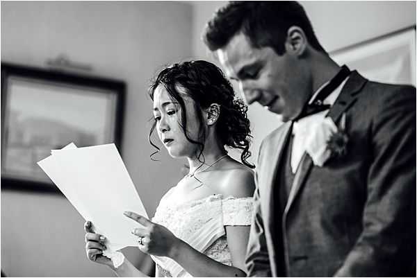 Black-and-white close-up portrait of a bride and groom during what appears to be a ceremony vow exchange indoors. The bride wears an off-the-shoulder lace dress with her dark hair in an updo, holding printed pages and reading aloud; her expression is focused and emotional. The groom stands beside her in a dark suit with a bow tie and a small floral boutonniere, also looking downward at the papers. The high-contrast monochrome tones give the image a sharp, documentary feel, with a framed artwork visible softly out of focus in the background.
