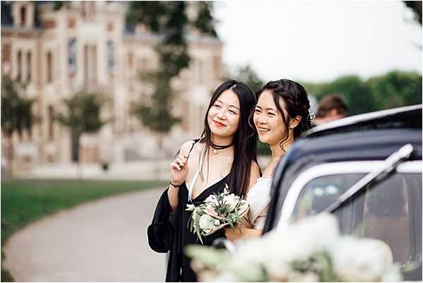 A bride and a female guest or bridesmaid pose together outdoors beside a dark-colored vintage car, with a large French chateau visible in the soft-focus background. The bride wears a white off-shoulder dress and holds a bouquet of white flowers with green eucalyptus foliage, while her companion wears a black outfit with a wrap or jacket. Both women are smiling and leaning close together in a candid, relaxed moment. The shot is a mid-range portrait with a shallow depth of field, taken on the chateau grounds along a gravel driveway.