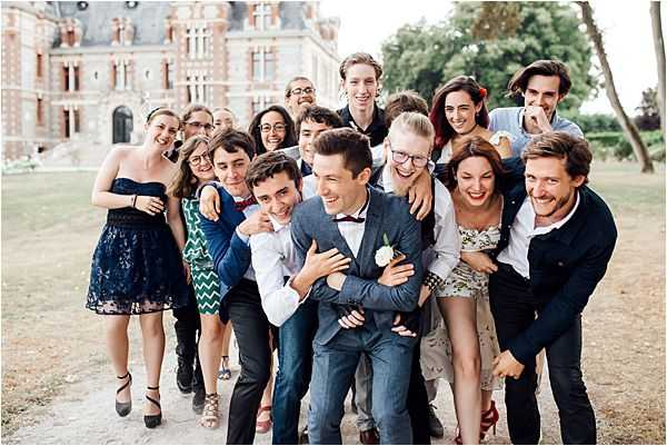 A group portrait taken outdoors on the grounds of a French chateau, showing approximately 15 guests playfully crowding around and embracing what appears to be the groom, who is wearing a navy blue suit with a burgundy bow tie and a white boutonniere. The guests are dressed in smart-casual wedding attire including a navy strapless floral mini dress, a teal geometric-print dress, and various suits and summer dresses in mixed colors. Everyone is laughing and smiling in a candid, energetic pose, with arms wrapped around each other in a jovial pile. The shot is taken at mid-distance, capturing the full group in a wide portrait orientation, with the red-brick chateau facade visible in the background. Potential venue feature image.