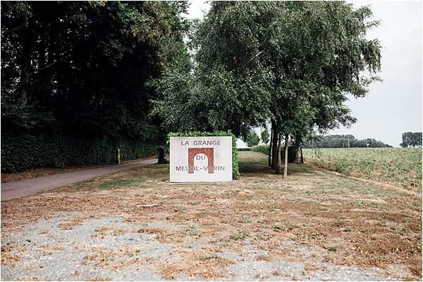 Wide shot of the entrance signage for La Grange du Mesnil-Varin, a French wedding venue. The sign is a painted white rectangular panel with red lettering and a red arch motif, positioned at a gravel driveway entrance flanked by mature trees and open fields. No people are visible in the frame. Potential venue feature image.