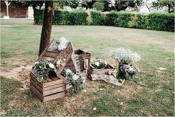 A rustic outdoor decor vignette photographed in a wide shot on a lawn, featuring a collection of weathered wooden crates stacked and arranged around the base of a tree. The crates are styled with small bouquets of white and blush flowers including baby's breath and what appear to be roses, along with burlap ribbon accents and sheer tulle fabric. Additional floral arrangements in zinc/metal buckets are placed nearby, continuing the white and green color palette. The overall styling theme is rustic-country, consistent with a French countryside wedding aesthetic.