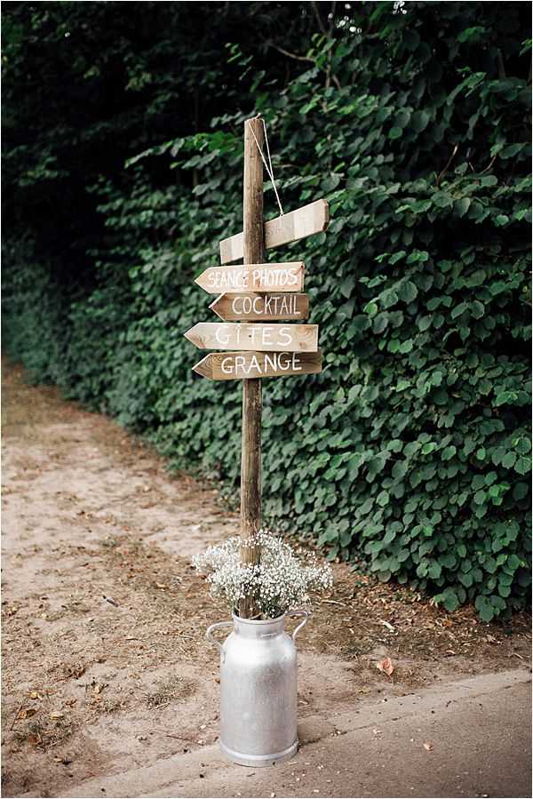 A rustic wedding directional signpost stands along a sandy path at an outdoor venue. The wooden post holds four hand-painted arrow signs in white lettering pointing guests toward 'Séance Photos,' 'Cocktail,' 'Gîtes,' and 'Grange.' At the base of the post, a silver milk churn serves as a vase holding a bouquet of white gypsophila (baby's breath). The overall styling is rustic-country, and the detail shot is taken at medium distance with a shallow depth of field.