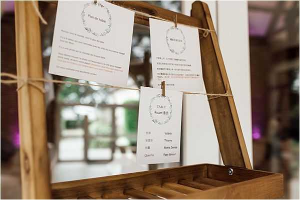 Close-up detail shot of a wedding seating chart display, featuring white cards with wreath illustrations hung by twine from a rustic wooden easel. The cards include a 'Plan de table' header card written in both French and Chinese, individual table assignment cards, and a visible table card labeled 'Table Rouen' with guest names listed. The bilingual signage suggests a Franco-Chinese wedding. The background is softly blurred, showing warm ambient lighting with hints of purple uplighting, indicating an indoor or covered reception venue with a rustic decor style.