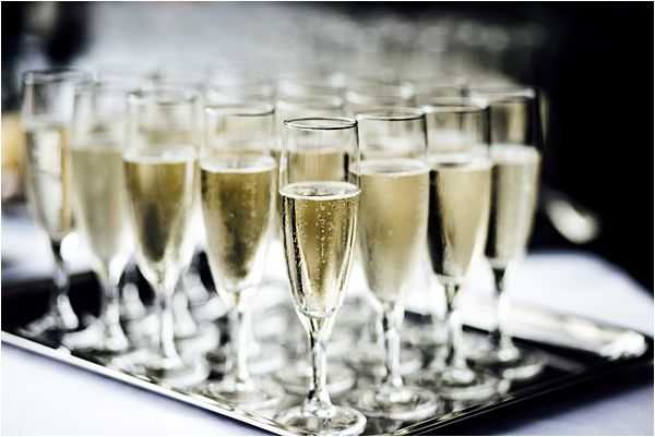 Close-up detail shot of approximately 20 champagne flutes filled with pale golden sparkling wine, arranged in rows on a polished silver serving tray, ready for a cocktail hour or toast service. The glasses are classic flute style, and the bubbles are visible in the liquid. The image has a shallow depth of field, with the front glasses in sharp focus and the back rows softly blurred. The background is out of focus with dark and white tones suggesting an indoor reception setting.