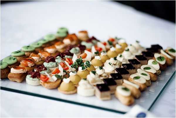 Close-up detail shot of a cocktail hour canapé display arranged on a glass serving tray, likely at a wedding reception. The tray features multiple rows of bite-sized hors d'oeuvres including smoked salmon on blinis with green herb cream, tomato and cream cheese crostini, olive and tapenade toasts, grape and cream cheese bites, dark chocolate and cream layered petit fours, and cucumber-topped rounds garnished with fresh herbs. The variety and uniform arrangement suggest formal French-style catering. The image is slightly shallow in depth of field, with the foreground canapés in sharp focus and the background softly blurred.