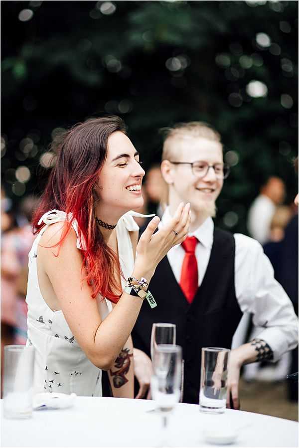 Two wedding guests are seated at a table during what appears to be an outdoor reception, laughing and reacting — likely during speeches or entertainment. The woman in the foreground has long red-dyed hair, a black choker necklace, a white sleeveless dress printed with small bird motifs, visible arm tattoos, and a festival-style wristband. The person beside her wears a white dress shirt, black vest, and a bright red tie, with short blonde hair and glasses. Water glasses are visible on the white-clothed table in the foreground. The setting is an outdoor garden with blurred greenery and other guests in the background. The overall styling is alternative and edgy rather than traditional.