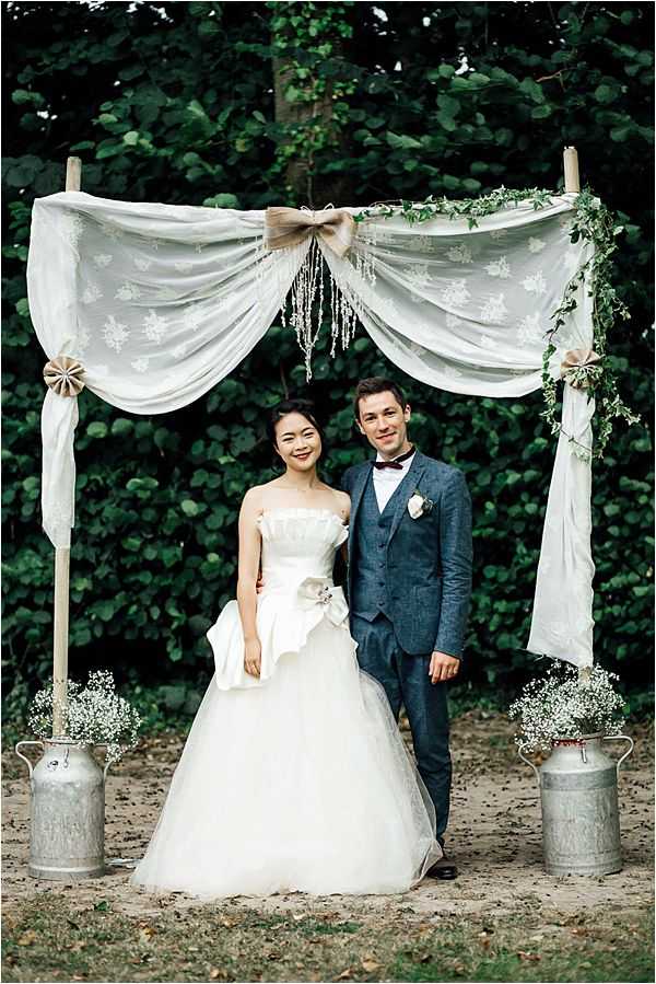 A couple poses together outdoors in front of a rustic ceremony arch made from wooden posts draped with ivory embroidered fabric, secured with burlap bows, and decorated with trailing green ivy and hanging crystal strands. Vintage galvanized milk churns holding white baby's breath arrangements flank the base of the arch on either side. The bride wears a strapless ivory ball gown with a large satin bow detail at the waist, while the groom wears a navy blue three-piece suit with a burgundy bow tie and a white boutonniere. The setting is an outdoor garden or wooded area, and the overall styling is rustic-romantic. Medium portrait shot of the two subjects framed centrally within the arch structure.