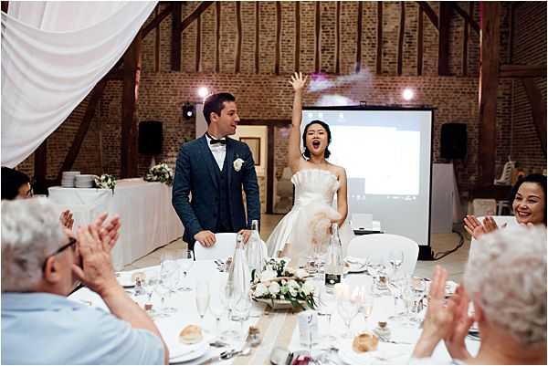 The bride and groom are making a celebratory entrance into the reception hall as seated guests applaud around them. The bride, wearing a strapless ivory ball gown, has one arm raised in excitement, while the groom wears a dark teal three-piece suit with a bow tie and white boutonniere. The setting is a rustic converted barn interior with exposed brick walls and wooden beamed ceiling, decorated with draped white fabric. Reception tables are set with white linens, crystal glassware, and low centerpieces of white roses and neutral-toned blooms. A projection screen is visible in the background. The image is a wide shot capturing the couple's joyful reaction alongside the enthusiastic response of approximately six visible guests in the foreground.