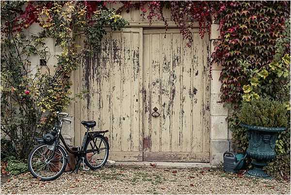 A wide shot of a rustic venue entrance featuring a pair of large, weathered cream-painted wooden doors framed by red and green climbing ivy in autumn foliage. A black bicycle is leaned against the wall to the left of the doors, and a dark green ornamental urn planter sits to the right alongside a watering can. The overall palette is muted and autumnal, with peeling paintwork on the doors adding a worn, characterful feel to the facade. No people are visible in the frame. Potential venue feature image.