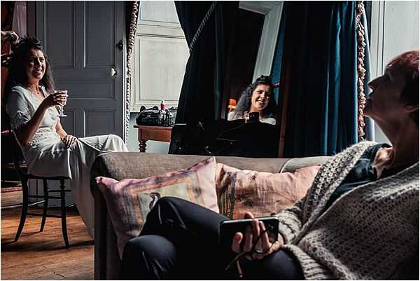 A candid getting-ready scene captured indoors in what appears to be a chateau or period-style room with wood flooring, paneled grey doors, and teal-blue curtains. The bride, wearing a white dress with her hair styled up, sits on a chair holding a champagne flute and laughing. In the foreground, a woman in casual clothes reclines on a grey sofa with pink and mauve linen cushions, holding a phone. A third woman is reflected in a large ornate mirror in the background, also laughing. The atmosphere is relaxed and informal, with the group clearly enjoying the pre-ceremony preparations. Medium-wide shot taken from a low angle.