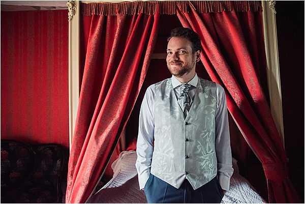 The groom is photographed getting ready indoors, standing in front of an ornate four-poster bed dressed with deep red damask curtains and gold fringe trim, suggesting a chateau or historic venue bedroom. He is wearing a light blue-grey damask waistcoat over a white dress shirt with a grey patterned cravat-style tie and navy blue trousers. He is smiling and looking slightly off-camera in a relaxed portrait pose. The rich red and gold backdrop contrasts with his silver-blue attire, reflecting a classic, formal wedding style.