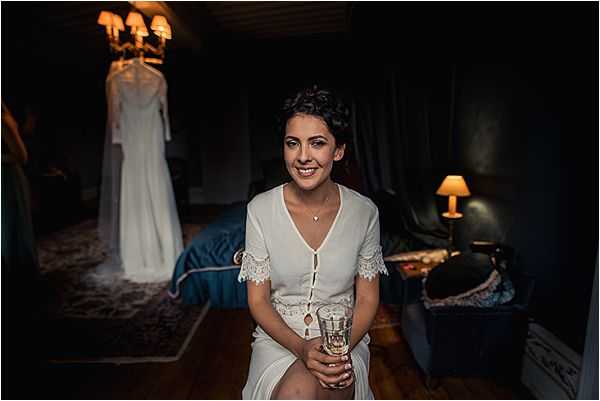 A bride sits in a dimly lit bedroom during the getting-ready phase, wearing a white short-sleeve dress with lace trim cuffs and holding a glass. Her dark hair is styled in an updo and she wears a delicate pendant necklace. In the background, her ivory wedding gown with long sleeves hangs on a hanger beneath a warm chandelier, while a teal bedspread and a small table lamp add to the moody, intimate atmosphere of the room. The portrait is a medium shot with shallow depth of field, emphasizing the bride in the foreground against the softly blurred bridal suite setting.