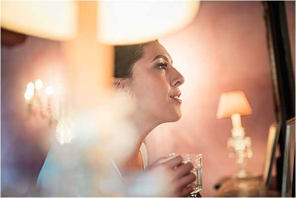 A close-up portrait of a bride during the getting-ready phase, captured in profile as she looks upward with a slight smile, holding a small glass vessel. The setting appears to be an indoor dressing room or bridal suite, warmly lit by table lamps and what appears to be a chandelier reflected in the background. The bride wears a white or ivory top and a delicate earring, with her dark hair styled up. The image has a soft, warm amber and pink tone created by the ambient lamp lighting, with intentionally blurred foreground elements — likely tulle or veil fabric — adding depth to the close-up composition.