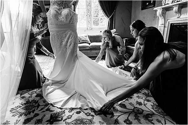 Black-and-white getting-ready scene shot indoors in what appears to be a ornate room with a decorative fireplace mantel, patterned carpet, and large windows with draped curtains. The bride stands at center wearing a fitted, lace-bodice wedding gown with a long smooth train, while three bridesmaids crouch and kneel around her to arrange and spread the train across the floor. A fourth person is visible at the left edge assisting with the veil. The image is captured in high-contrast black and white with deep shadows, and is composed as a wide shot slightly from below, emphasizing the scale of the dress and the collaborative effort of the bridal party.