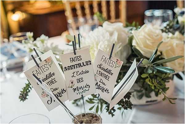 A close-up detail shot of a wedding table centerpiece featuring several small sparkler packets on wire stems, each printed with the French phrase 'Faites Scintiller L'Entrée des Mariés' ('Sparkle the Entrance of the Newlyweds'), decorated with small floral illustrations in blush tones. The sparkler packets are arranged in a small pot surrounded by greenery, with a large floral arrangement of ivory roses and eucalyptus visible in the background. The table is set with white linens and glassware, and the reception venue interior is softly lit and warm-toned in the background. The overall decor palette is white, green, and blush, consistent with a classic romantic French wedding reception styling.
