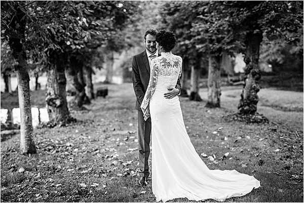 Black-and-white couple portrait taken outdoors in a tree-lined avenue, with the trees receding into the background to create a strong sense of depth. The bride faces away from the camera, revealing the back of her long-sleeved lace wedding gown with a flowing train, while the groom faces toward the camera smiling, dressed in a dark suit. The two are embracing, with the bride's arms around the groom's waist. The image is shot in medium-wide format with high contrast black-and-white tones, emphasizing the texture of the lace dress and the dappled light filtering through the tree canopy.