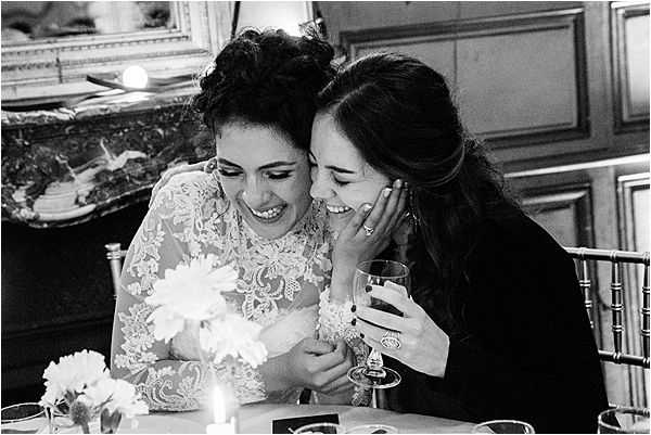 Black-and-white close-up portrait of a bride and a female guest sharing a laugh at a reception table inside what appears to be a chateau or formal ballroom, with an ornate fireplace mantel visible in the background. The bride wears a long-sleeve lace gown with floral appliqué detailing and has her dark hair pinned up, while her companion holds a wine glass and leans in closely. A lit candle, scattered flower petals or small blooms, and what appear to be printed photographs sit on the table in front of them. The image has warm mid-tones and soft contrast, capturing an candid, joyful moment between the two women.