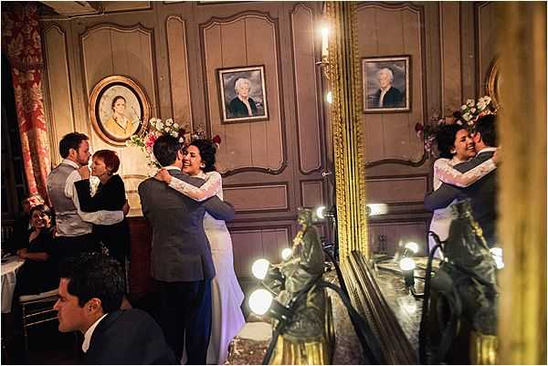 A first dance scene captured indoors in what appears to be a classic French chateau or manor room, with wood-paneled walls painted in a warm taupe and framed portrait paintings hanging on the wall. The bride wears a white gown with lace long sleeves and the groom is dressed in a dark navy suit; they are embracing and smiling during their first dance. A large ornate gold-framed mirror to the right reflects the couple, creating a dual view of the moment, while warm Edison-style bulb lighting along the mirror frame adds a soft glow. In the background, two other guests — one in a light grey suit — are also embracing or dancing, and a seated male guest in a dark suit is visible in the foreground.