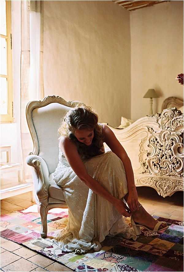 A bride is getting ready, seated in a white ornate French Louis XVI-style armchair as she fastens her shoes. She is wearing a fitted, ivory sequined or beaded gown and is looking down at her feet. The room features a heavily carved white Baroque-style bed frame, a small bedside lamp, warm plaster walls, and natural light streaming through a tall window with sheer curtains. A colorful patchwork rug in pastel tones of pink, purple, blue, and green lies beneath her on terracotta tile flooring. The overall styling is romantic and vintage-inspired. Medium portrait shot taken from a slight distance.