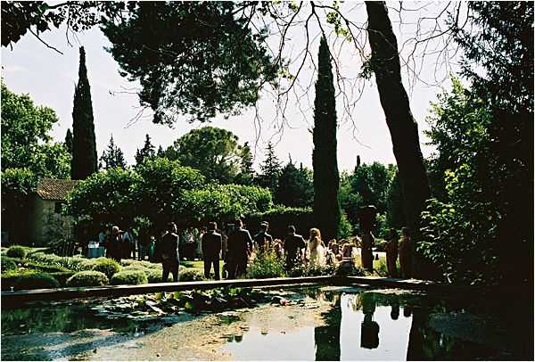 An outdoor wedding ceremony taking place in a formal garden setting, with approximately 20–30 guests gathered around what appears to be a ceremony area among sculpted hedges and tall cypress trees. The shot is taken from behind a long rectangular reflecting pool or ornamental pond in the foreground, with lily pads visible on the water surface and the ceremony scene mirrored in the reflection. The figures are largely silhouetted against bright afternoon backlighting, making individual details difficult to distinguish, though a bride in a white dress is faintly visible among the group. Wide-angle shot capturing the full garden landscape and water feature.