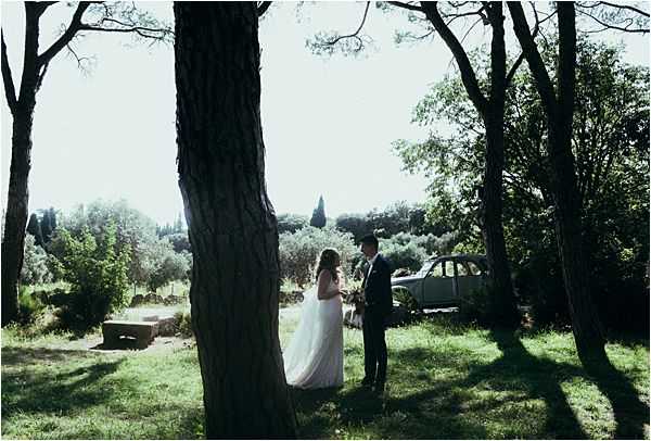 A couple shares a quiet moment together in an outdoor wooded setting, standing face to face among tall trees on a grassy lawn. The bride wears a white floor-length gown and holds a bouquet, while the groom is dressed in a dark suit. A vintage pale green/grey classic car is parked in the background, adding a retro styling element to the scene. The image is a wide shot with strong backlighting that creates silhouette-like contrast between the dark tree trunks and the bright, overexposed sky beyond.