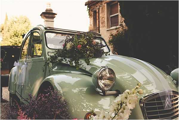 A detail shot of a sage green vintage Citroën 2CV decorated as a wedding car, photographed outdoors in front of a stone building. The hood of the car is adorned with a loose floral arrangement featuring deep burgundy and pink blooms with trailing greenery, while a garland of white flowers — appearing to be white delphinium or similar tubular blooms — drapes across the front grille and bumper. The styling is rustic and romantic, consistent with a French countryside or Provençal wedding aesthetic. The image is a close-up, slightly warm-toned shot emphasizing the floral decorations and the car's curves.