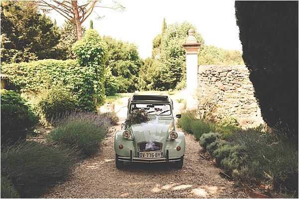 A pale mint-green vintage Citroën 2CV is parked on a gravel driveway, decorated with a floral arrangement of greenery and blooms on its hood, serving as a wedding car. The setting is an outdoor estate entrance with a stone pillar gateway, dry-stone walls, and lavender plants lining the driveway. No people are visible in the frame. The composition is a straight-on wide shot, giving the scene a classic French countryside aesthetic. Potential venue feature image.