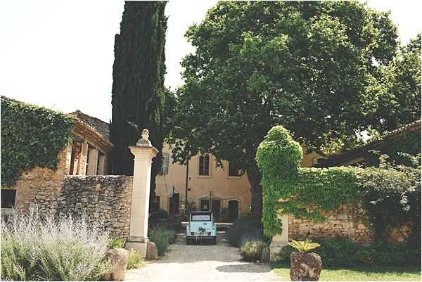 Wide exterior shot of a French country estate or mas, viewed through stone entrance pillars topped with classical busts. A vintage pale blue Citroën 2CV or similar retro vehicle is parked on the gravel driveway leading up to the main ochre-yellow stone building. The property features low stone outbuildings covered in climbing ivy on either side of the entrance, with cypress trees and a large shade tree visible in the courtyard. No people are visible in the frame; the shot appears to document the venue and its vintage wedding car styling detail. Potential venue feature image.