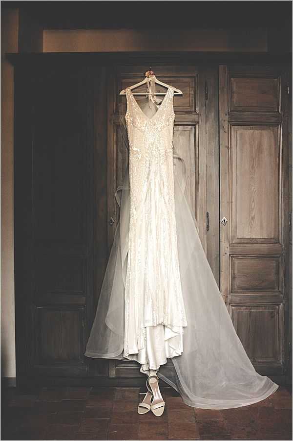 A getting-ready detail shot showing an ivory, heavily beaded and lace-embroidered sleeveless wedding gown hanging on a wooden hanger against a set of large, dark wood panelled doors. The fitted, V-neck dress features intricate floral beading across the bodice and skirt, with a long sheer tulle veil or train draping to the floor on both sides. A pair of white strappy block-heel sandals is placed on the terracotta tile floor directly beneath the gown. The warm, dim interior lighting creates a dark, moody atmosphere that contrasts with the brightness of the dress. The composition is a wide vertical portrait shot centered on the dress.