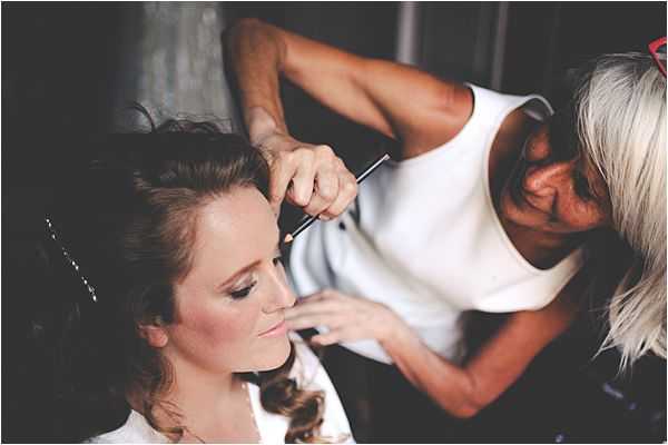 A getting-ready scene showing a bride having her makeup applied by a makeup artist. The bride is seated, wearing a white robe, with her hair styled in loose curls and secured with small decorative pins. The makeup artist, dressed in a sleeveless white top with short silver-grey hair, is applying eye makeup using a fine brush. The image is a close-up portrait shot with a dark, moody background and soft natural lighting falling on the bride's face.