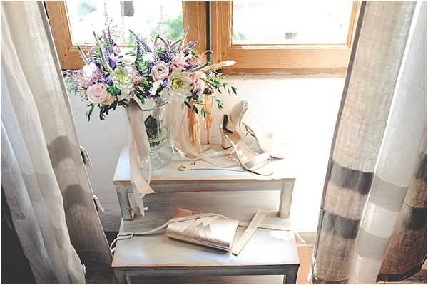 A detail flat-lay style shot of bridal accessories arranged on a small white wooden step stool near a window with sheer linen curtains. The bouquet features lavender, soft lilac, blush pink, and white blooms — including what appear to be ranunculus, sweet peas, and delphinium — with trailing ivory satin ribbons, displayed in a glass vase. A pair of strappy nude/champagne heeled sandals and a rose gold or blush satin clutch bag are styled alongside the bouquet. The overall color palette is soft and romantic, combining blush, champagne, and lavender tones in a light-filled indoor setting.