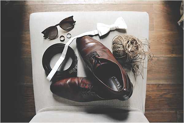 A flat lay detail shot of groom's wedding accessories arranged on a light grey upholstered chair seat. Items include a pair of dark brown leather oxford dress shoes, a white bow tie, a black leather belt, a pair of dark wayfarers-style sunglasses, two gold wedding bands, and a small ball of natural twine. The composition is shot from directly above on a wooden floor background, with a rustic styling aesthetic suggested by the twine detail alongside the classic accessories.