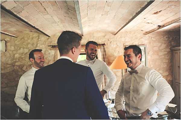 Four men are laughing together during what appears to be a getting-ready moment, photographed indoors in a room with exposed stone walls and a vaulted ceiling typical of a French farmhouse or chateau. The groom, seen from behind, is wearing a navy suit, while the three groomsmen are dressed in white dress shirts — one of whom is wearing a blush pink bow tie. A warm-toned table lamp is visible in the background along with a window. The shot is a candid mid-range portrait capturing a relaxed, jovial atmosphere among the group.