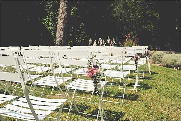 An outdoor wedding ceremony setup photographed before guests arrive, showing rows of white wooden folding chairs arranged on a lawn with a central aisle. Small floral arrangements in pink and deep burgundy tones are attached to the aisle-facing chairs as pew markers. The setting appears to be a garden or park with mature trees in the background. Wide shot with a slightly soft, film-like quality to the image.
