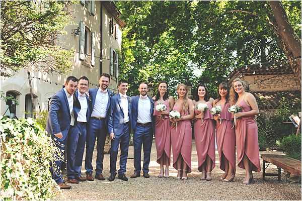 A bridal party group portrait taken outdoors in the courtyard of a French country property, likely a mas or small chateau, with a rendered stone building visible in the background. The group of approximately nine people includes four groomsmen and the groom dressed in navy blue suits with white dress shirts, and four bridesmaids plus the bride arranged to the right, all wearing dusky rose draped midi dresses with nude heels. The bridesmaids carry small rounded bouquets of white flowers with greenery. The shot is a wide, evenly lit daytime portrait with the full group standing in a relaxed line on a gravel surface.