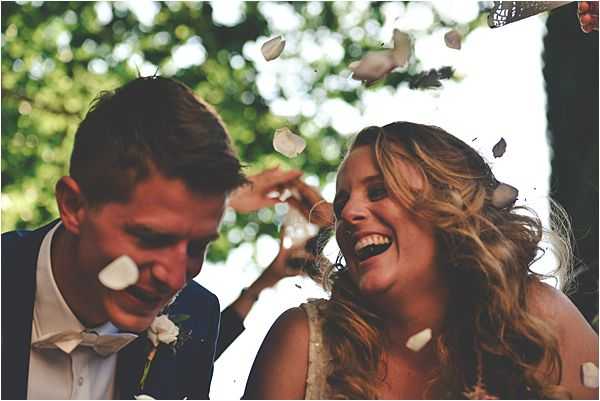 A close-up portrait of a bride and groom being showered with white flower petals, likely immediately after an outdoor ceremony. The groom wears a navy suit jacket with a white shirt and a small white floral boutonniere, while the bride wears a sleeveless dress and has wavy blonde hair. Both are laughing, with the bride's expression particularly joyful as petals land on and around them. The background is blurred green foliage, and the image has a warm, natural light quality.