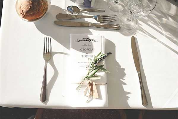 A close-up flat lay of a wedding reception table place setting shot from above in bright natural light. The white linen tablecloth holds a printed wedding dinner menu card reading 'Georgia & Florent,' tied with a thin ribbon and decorated with a small sprig of olive branch and a luggage-style tag. The setting includes polished silver cutlery — a fork on the left, a knife on the right, and additional cutlery and crystal wine glasses visible in the upper portion of the frame. A bread roll on a small plate is partially visible in the upper left corner. The overall styling is minimal and classic, with a greenery accent in keeping with a natural, understated aesthetic.