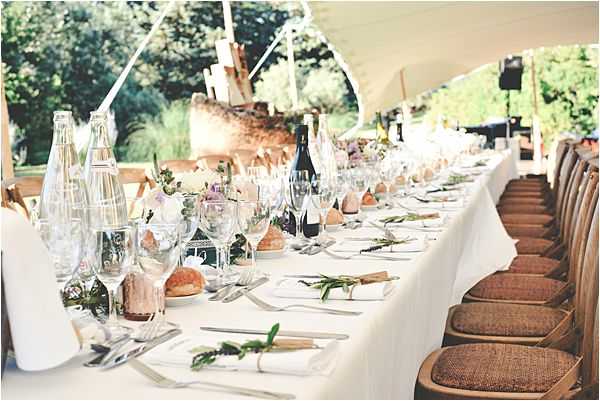 A wide shot of a long rectangular reception dining table set under an open-sided white marquee tent, photographed from one end looking down its length. The table is covered in a white linen cloth and set with crystal wine glasses, water carafes, silver cutlery, white napkins tied with sprigs of greenery, individual bread rolls, and small floral centerpieces featuring white and blush blooms with soft purple accents. Wine bottles and candle holders are spaced along the table. Brown wicker-seated wooden chairs line one side. The styling is relaxed and rustic, with a garden setting visible through the open tent sides in the background.