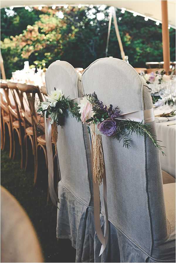 Detail shot of wedding reception seating under a white tent, showing the backs of chairs dressed in loose grey-linen slipcovers and decorated with floral posies featuring blush roses, purple/mauve garden roses, lavender, white blooms, and trailing rosemary sprigs, tied with ivory satin ribbon streamers and wheat-colored pampas-style tassels. The chairs are positioned along a long banquet table set with glassware and white linens, with natural wood cross-back chairs visible along the opposite side of the table. String lights are visible overhead inside the tent, and the overall styling is rustic-romantic with a soft purple, blush, and grey palette.