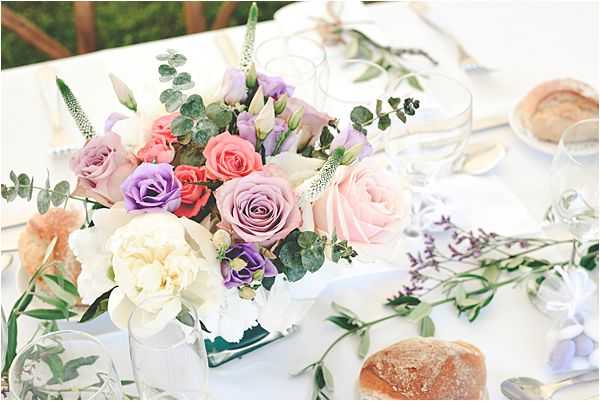 Close-up detail shot of an outdoor wedding reception table set in a garden setting. The centerpiece is a compact square vase arrangement featuring blush pink, coral, lavender, and purple roses alongside ivory peonies, purple lisianthus, veronica spikes, and eucalyptus foliage. The white linen table is styled with scattered eucalyptus branches, clear glassware, gold cutlery, folded white napkins, and individual bread rolls placed directly on the table. The overall decor palette is soft pastel with a romantic garden style.