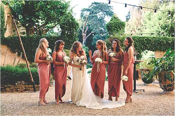 A bride and five bridesmaids stand together in a gravel courtyard, laughing and interacting with one another in a candid group portrait. The bride wears a lace, fitted ivory gown with a flowing train, while all five bridesmaids wear matching terracotta-rose wrap-style midi dresses with thin straps and nude heeled sandals. Each bridesmaid holds a small bouquet of white flowers, while the bride holds a larger white bouquet that appears to include white dahlias or peonies with minimal greenery. The outdoor setting features a hedge backdrop, festoon string lights strung overhead, and a rustic wooden A-frame structure to the left, suggesting a boho-rustic styling theme. The shot is a medium-wide portrait taken at eye level.